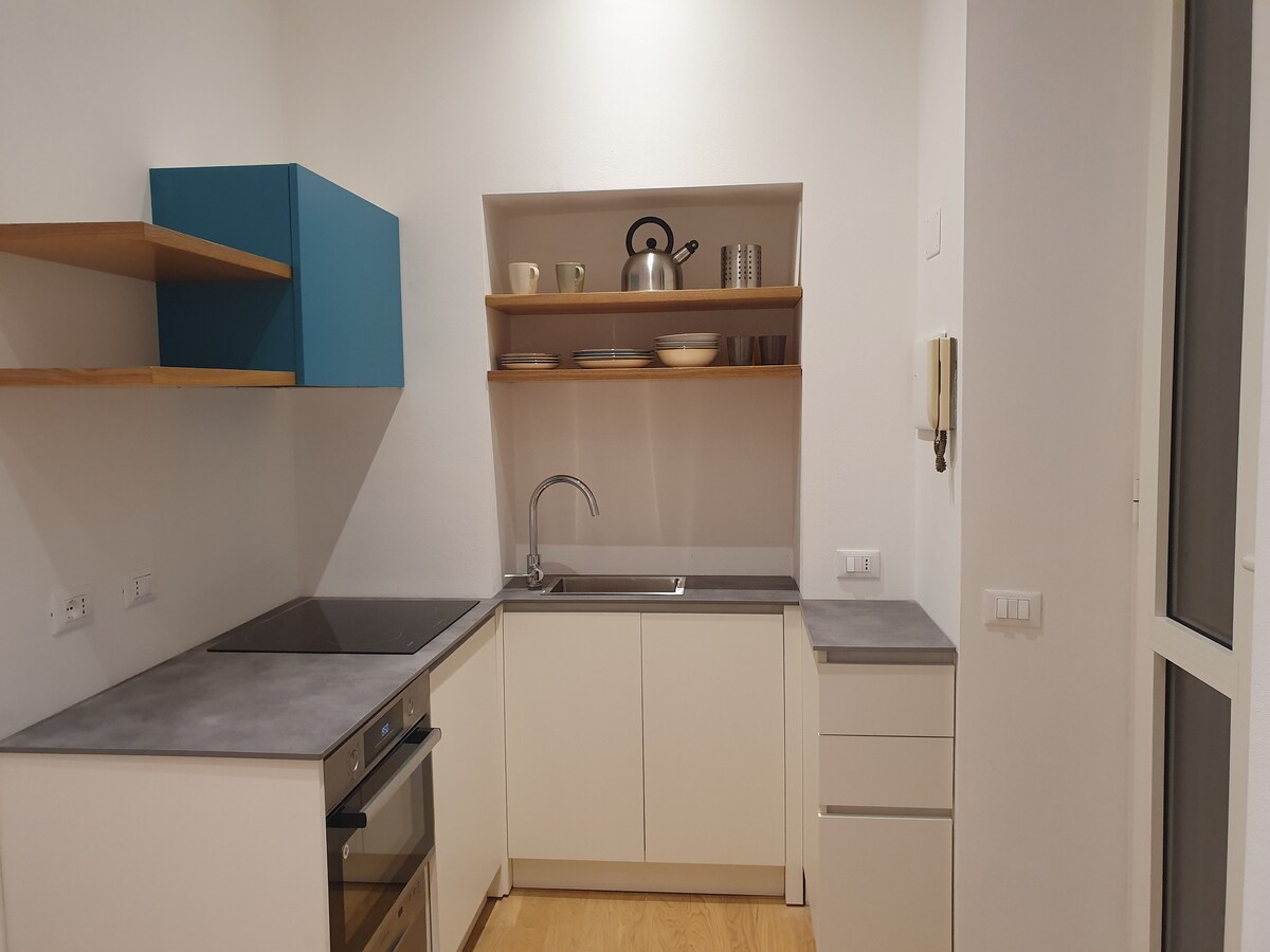 A modern kitchen area features minimalist cabinetry with open shelving. A sleek countertop integrates a stove and a stainless-steel sink, complemented by organized dishware and cooking utensils displayed above. Natural light enters through a nearby window, enhancing the bright atmosphere.