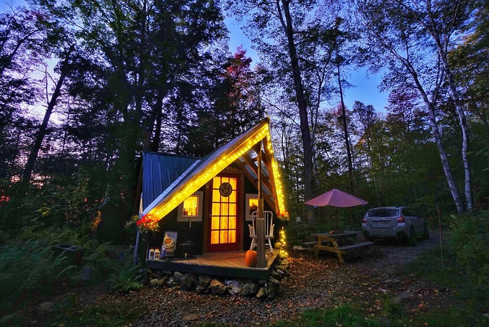 A Nordic inspired cabin is nestled among trees at dusk, with warm lights highlighting its unique triangular architecture. A picnic table and a vehicle are visible nearby, while the soft glow from the cabin enhances the surrounding natural beauty.