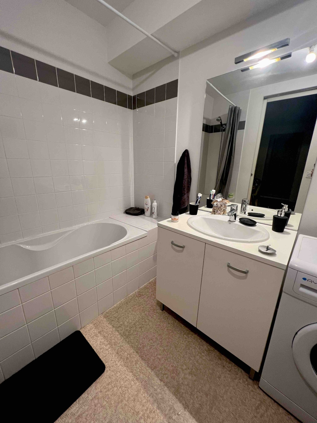 A clean bathroom is featured, showcasing a white bathtub alongside a dual sink vanity. A large mirror reflects the space, complemented by modern lighting. A washing machine is positioned in the corner, and towels are neatly hung nearby for convenience.