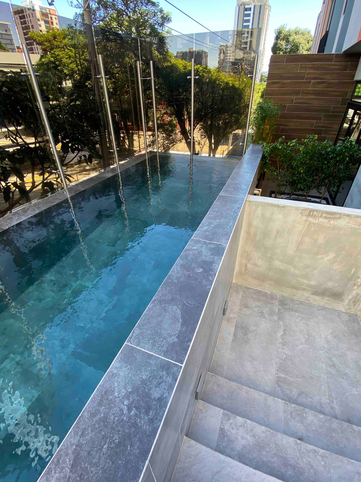 A modern climatized pool is nestled on a terrace, featuring transparent glass railings that provide an unobstructed view of the surrounding greenery. Stone steps lead down from the pool area, complementing the sleek design with light gray tiles.