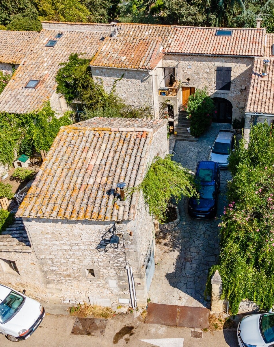 An aerial view captures the stone building's rustic charm, showcasing a terracotta-tiled roof and a peaceful courtyard lined with greenery. Several cars are parked along the stone-paved pathway, providing a sense of accessibility and ease in the tranquil village setting.