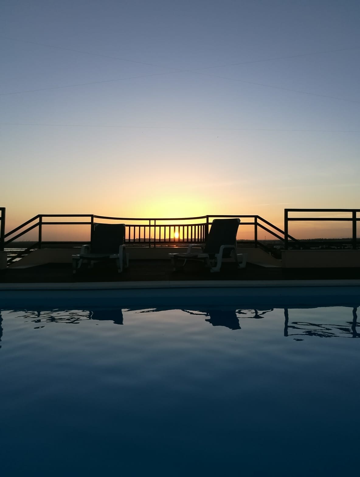 A tranquil scene captures the silhouette of lounge chairs against a sunset sky, reflecting golden hues on the calm surface of the pool. A railing surrounds the terrace, creating a serene atmosphere for relaxation.