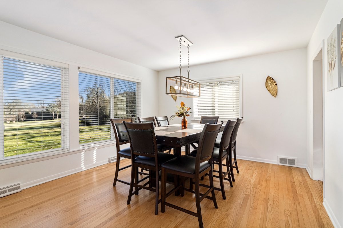 A bright dining area features a large dark wood table surrounded by eight black chairs. Natural light floods in through two expansive windows, offering views of the green outdoor space. A modern light fixture hangs above the table, adding a contemporary touch.