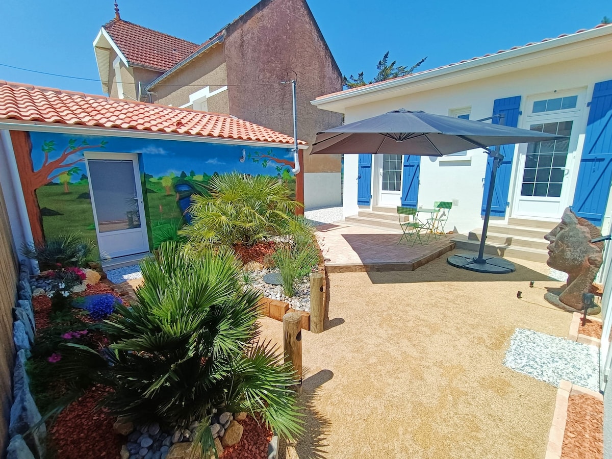 A colorful outdoor area is portrayed, featuring vibrant landscaping with palm plants and flowers. A shaded patio space is furnished with a table and chairs. The adjacent building displays a mural depicting nature, with blue shutters complementing the sunny ambiance.