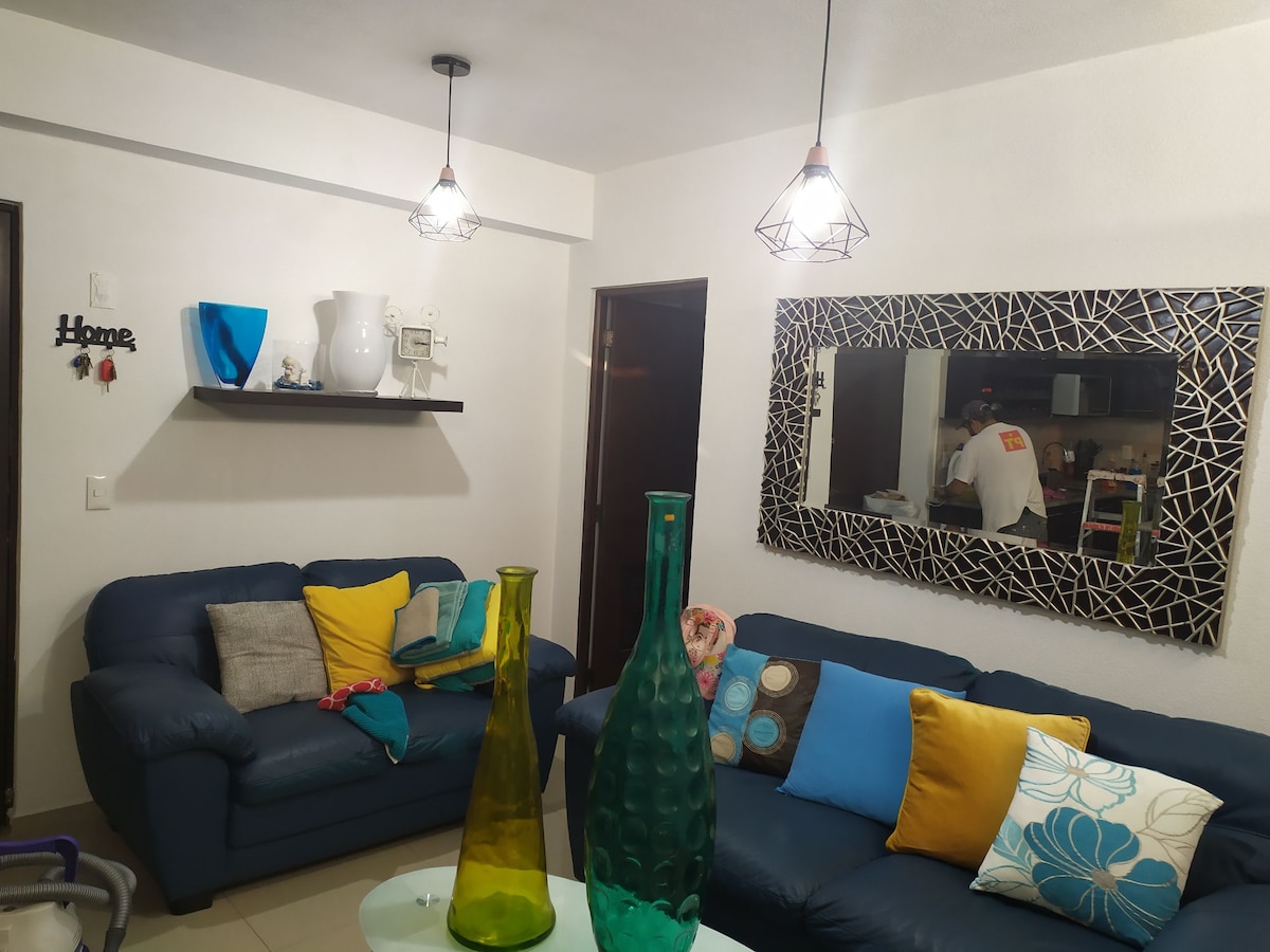 A cozy living area is featured, showcasing a dark blue sofa adorned with colorful cushions in yellow and turquoise. Two decorative glass vases in green and amber add a vibrant touch. Bright pendant lights hang from the ceiling, complementing the modern mirror on the wall.