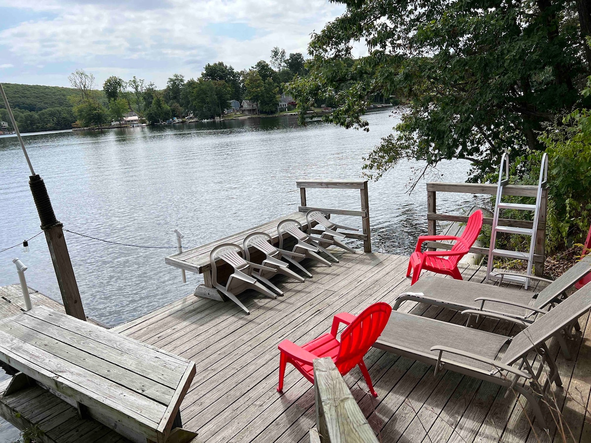 A lakeside deck is showcased with several lounge chairs arranged for relaxation. Light red chairs contrast with the wooden deck, leading to a calm lake view surrounded by trees. A ladder is visible, providing access to the water, enhancing the serene lakeside atmosphere.