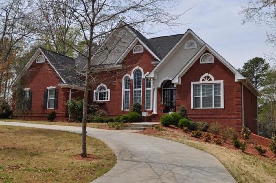 A spacious brick exterior of a single-story home is featured, surrounded by well-maintained landscaping. A curved driveway leads to the entrance, which includes multiple large windows and a wide front porch. Trees and shrubs frame the property, contributing to its peaceful setting.