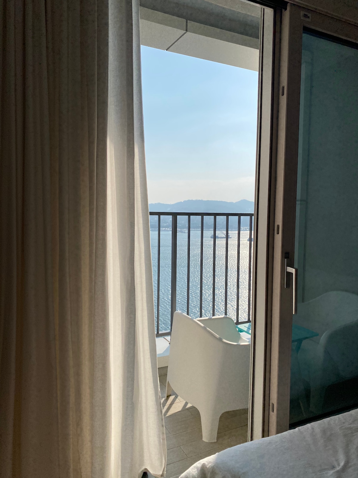 A serene view of the water is framed by sheer curtains and a glass door. A balcony with a chair is visible outside, allowing for a peaceful connection to the natural landscape beyond. The soft light reflects off the water, creating a calming ambiance.