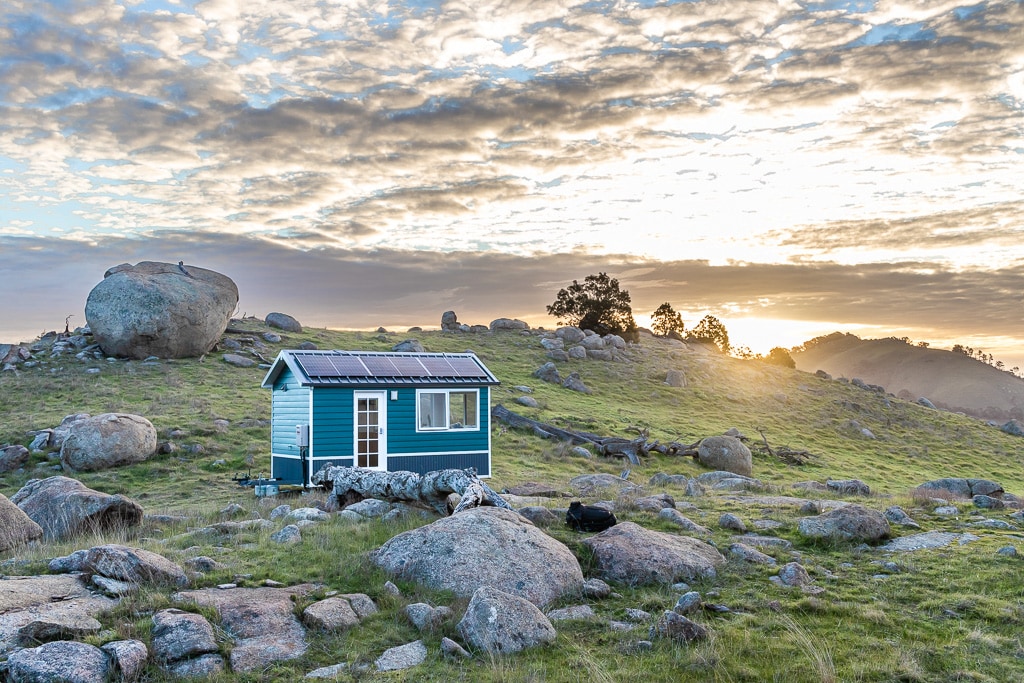 A tiny house is positioned on a grassy hill surrounded by large boulders, with solar panels mounted on the roof. The sky is filled with soft clouds, illuminated by a warm sunrise. The landscape features gentle hills in the background.