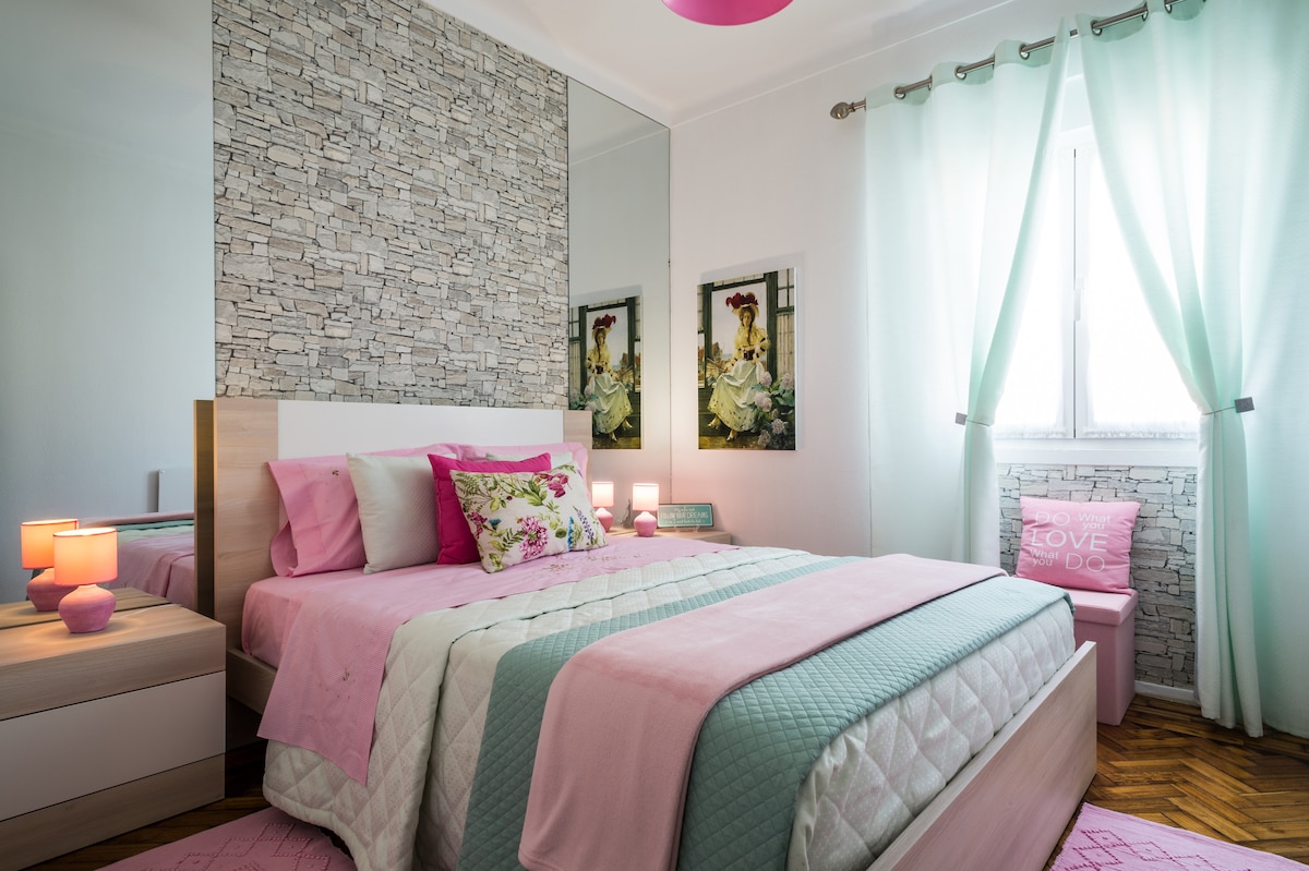 A well-lit bedroom features a spacious bed with pastel bedding and decorative pillows. Soft green curtains frame the window, while two framed artworks are displayed on the wall. A bedside table with a lamp and a small stool adds functionality and charm.