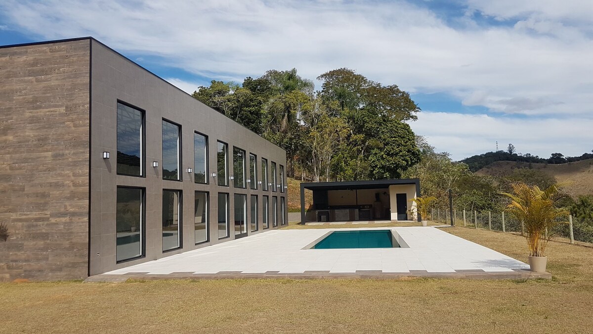 A modern building with large glass windows is set against a backdrop of trees and hills. A rectangular pool is surrounded by a spacious patio, creating an open area for relaxation. The landscape features well-maintained grass and a distant view of rolling terrain.