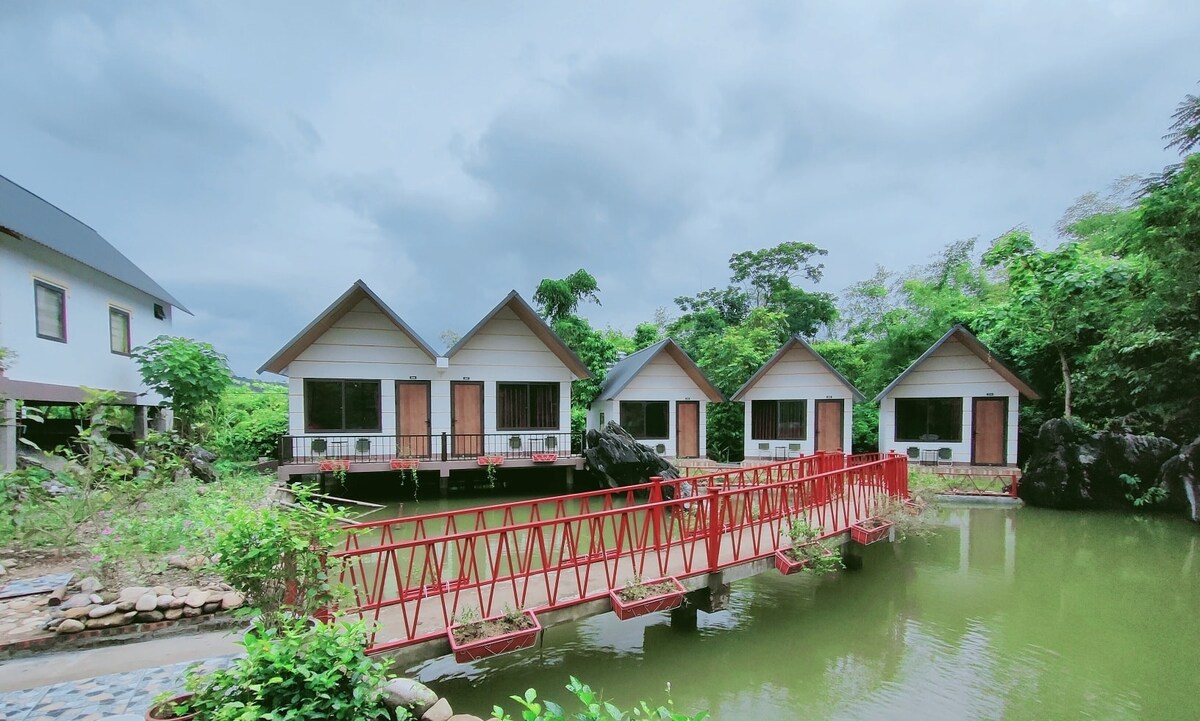 Four bungalows are situated on a calm pond, accessible via a red bridge. Each bungalow features an angled roof and large windows, surrounded by lush greenery. A tranquil atmosphere is created by the natural setting and reflective water.