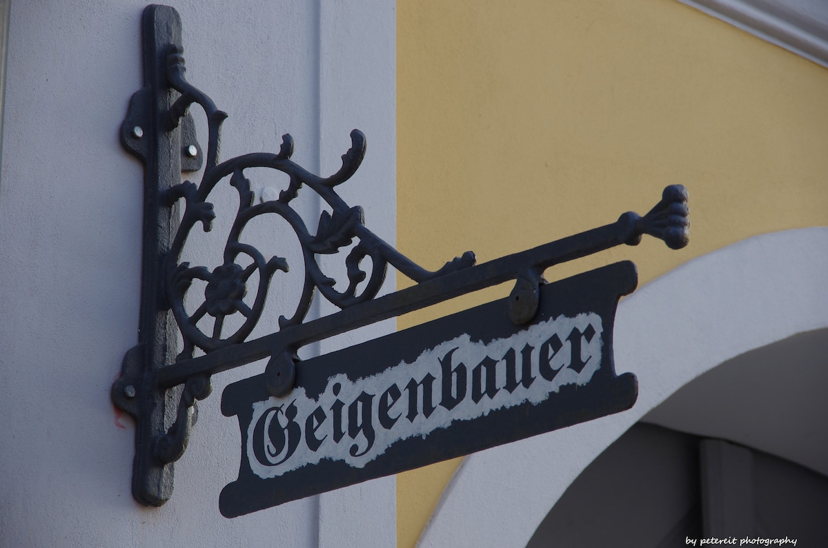 An ornate metal sign hangs from a decorative bracket, displaying the word 'Geigenbauer' in a classic font. The sign is mounted against a yellow wall, emphasizing the craftsmanship and historical charm of the location.