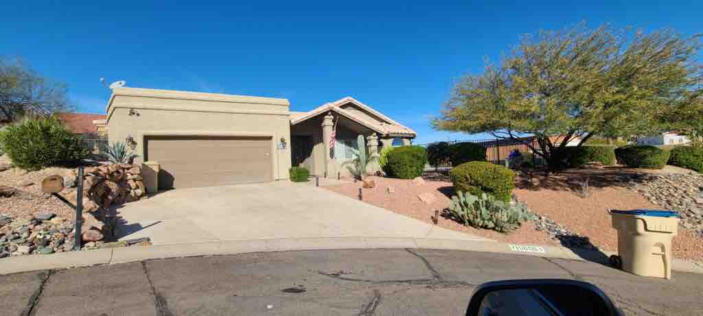 The exterior of a single-story home is presented, featuring a light-colored stucco facade and a tiled roof. A landscaped front yard includes various shrubs and cacti, complemented by a driveway leading to a two-car garage. A clear blue sky provides a bright background.