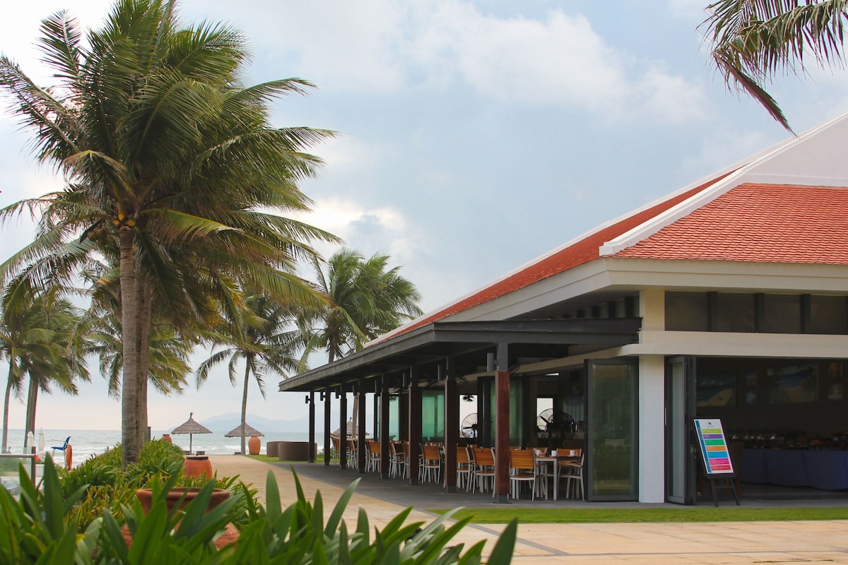 The beachfront restaurant features a spacious outdoor dining area surrounded by palm trees and well-kept landscaping. Large glass doors provide views of the ocean, with a partially cloudy sky in the background. Umbrellas and a distant beach can be seen through the trees.
