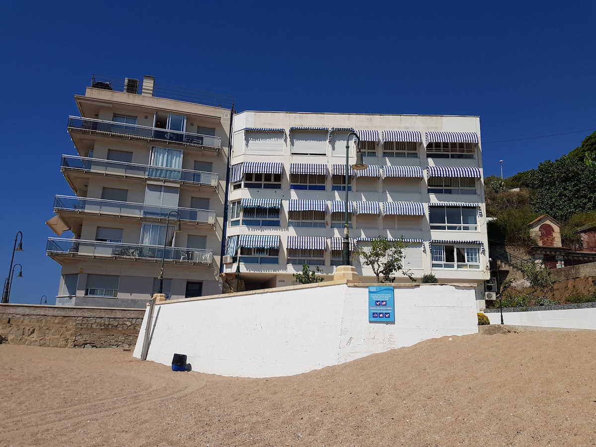 The building features multiple balconies adorned with striped awnings, overlooking a sandy beach. The structure is composed of several stories, with large windows allowing for ample natural light. A clear blue sky and the beach ambiance create a tranquil seaside setting.