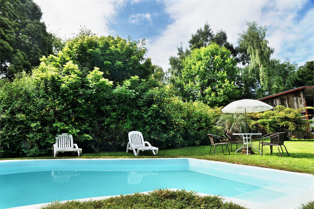 A serene outdoor space features a clear swimming pool surrounded by lush greenery. Nearby, a round table with chairs is shaded by an umbrella, and two white lounge chairs are positioned on the grass, inviting relaxation.