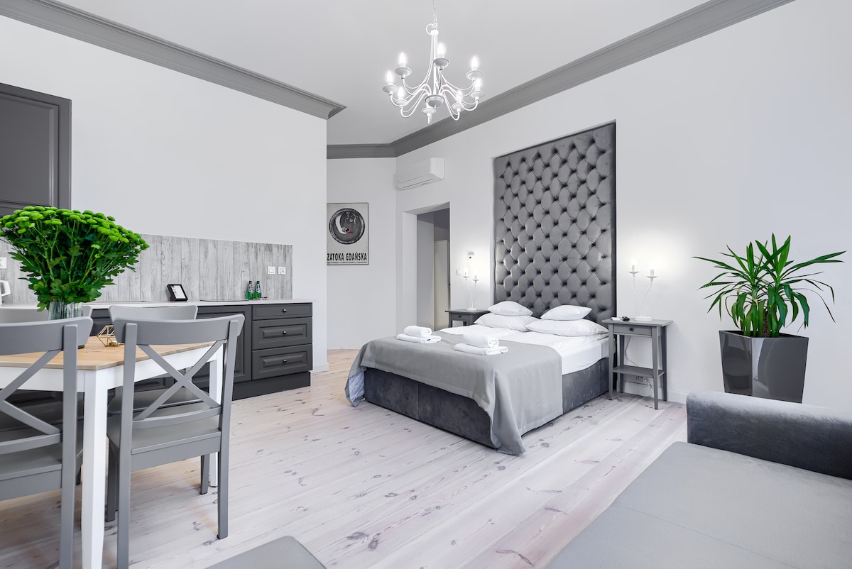 A spacious studio presents a comfortable double bed with a tufted headboard. A dining table is set for two, complemented by modern decor. Soft lighting from the chandelier enhances the bright atmosphere, while plants add a touch of greenery to the inviting space.