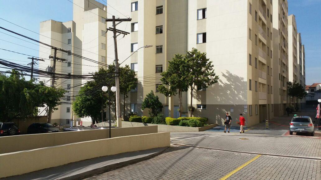 The exterior of a multistory residential building is shown, featuring light-colored façades and a landscaped area. Paved pathways and parked cars are visible, along with electric poles and trees providing greenery. Two individuals walk alongside the building, contributing to the community atmosphere.