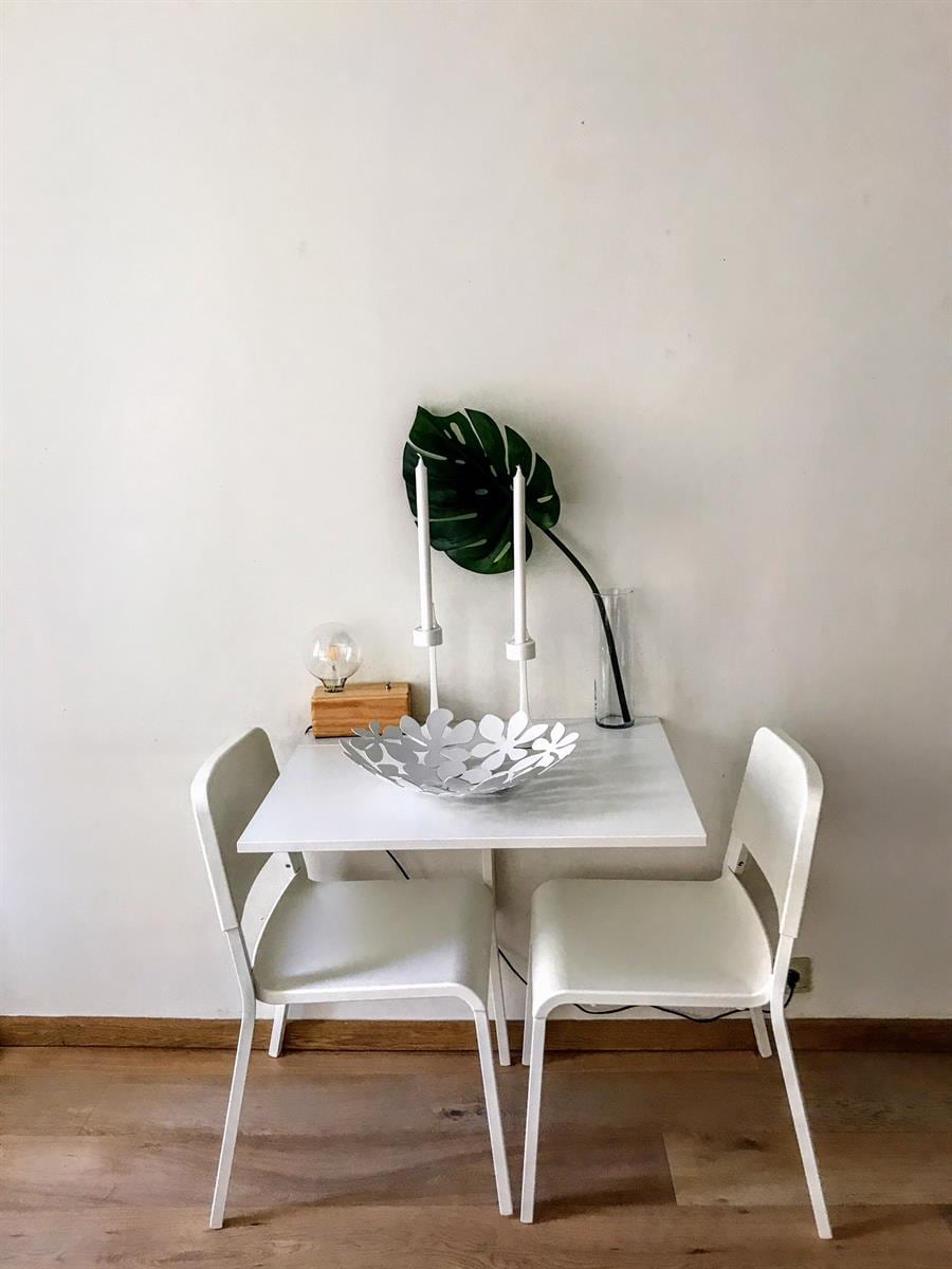 A small dining area features a white table paired with two chairs. A decorative bowl sits on the table, complemented by a green leaf placed in a clear vase. A candle holder with three candles adds a subtle touch of elegance.