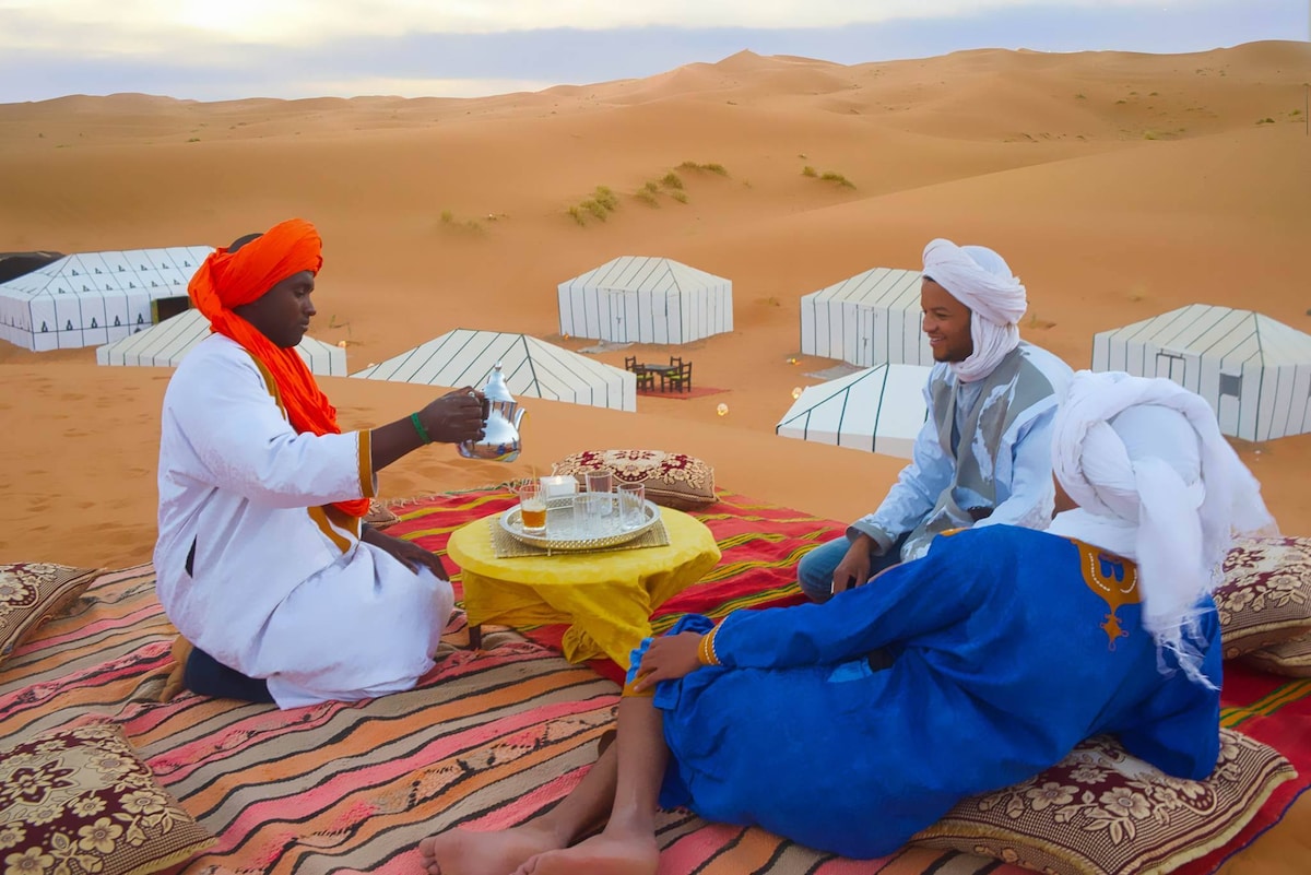 A welcoming setting is seen in the desert, featuring traditional Moroccan tents in the background. A table with tea is being prepared, while individuals in colorful attire sit on patterned textiles. The expansive sandy dunes create a serene backdrop under a cloudy sky.