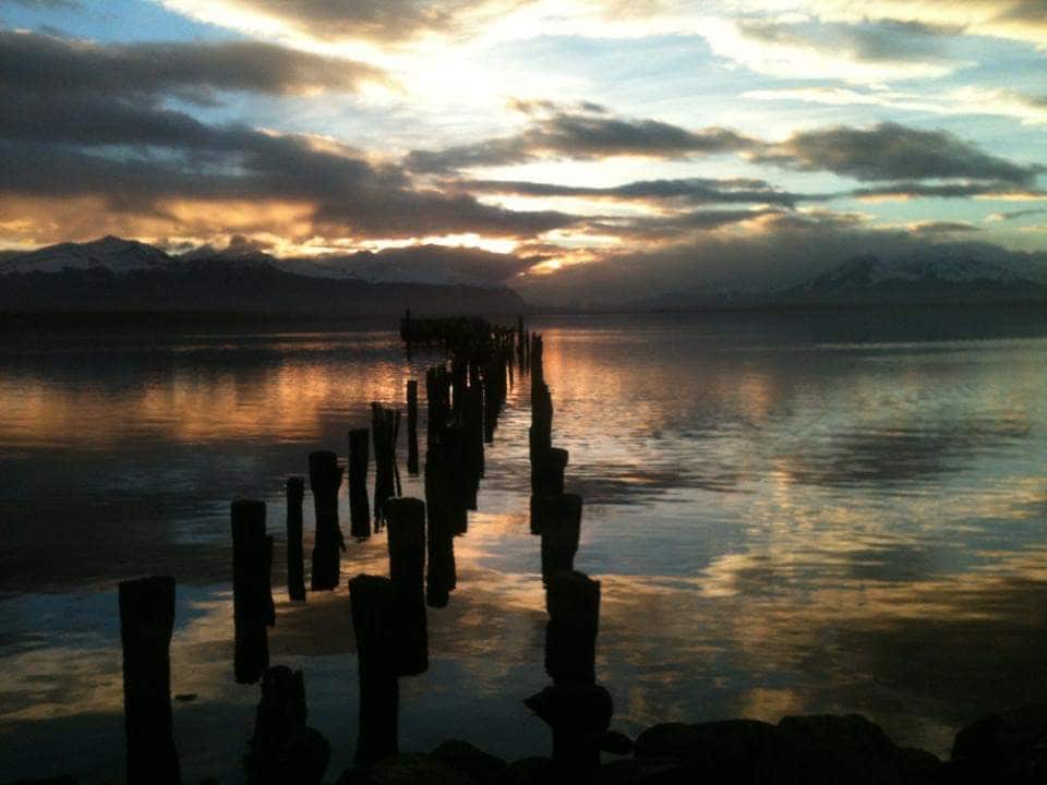 A weathered wooden pier extends into calm waters, creating a pathway towards the horizon. The surface reflects vibrant colors from the sky, with clouds softly illuminated by the setting sun. Majestic mountains can be seen in the background, adding depth to the serene landscape.