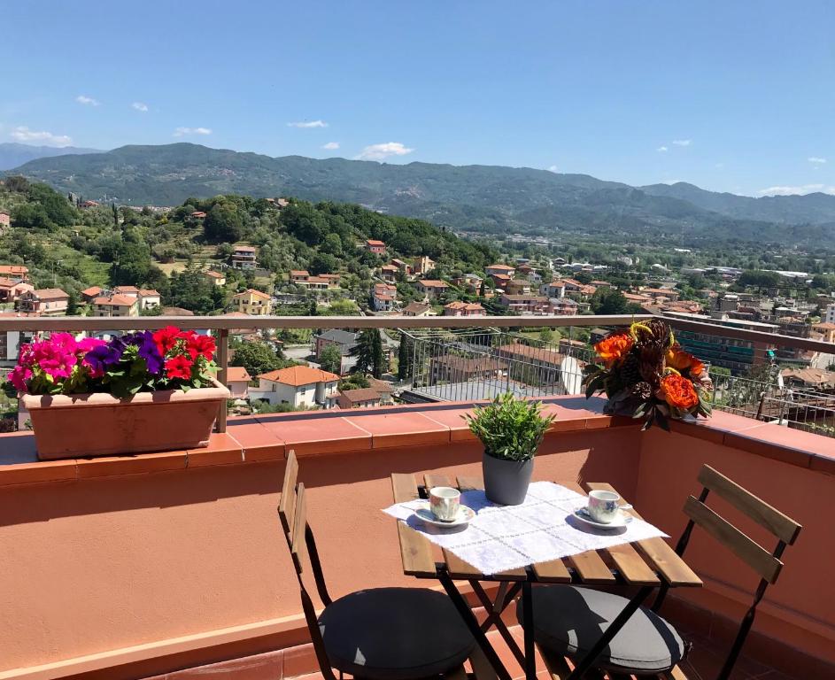 A spacious terrace is adorned with blooming flowers in planters, offering a table set for two. The surrounding scenery reveals rolling hills and a valley, with a clear blue sky above enhancing the view.