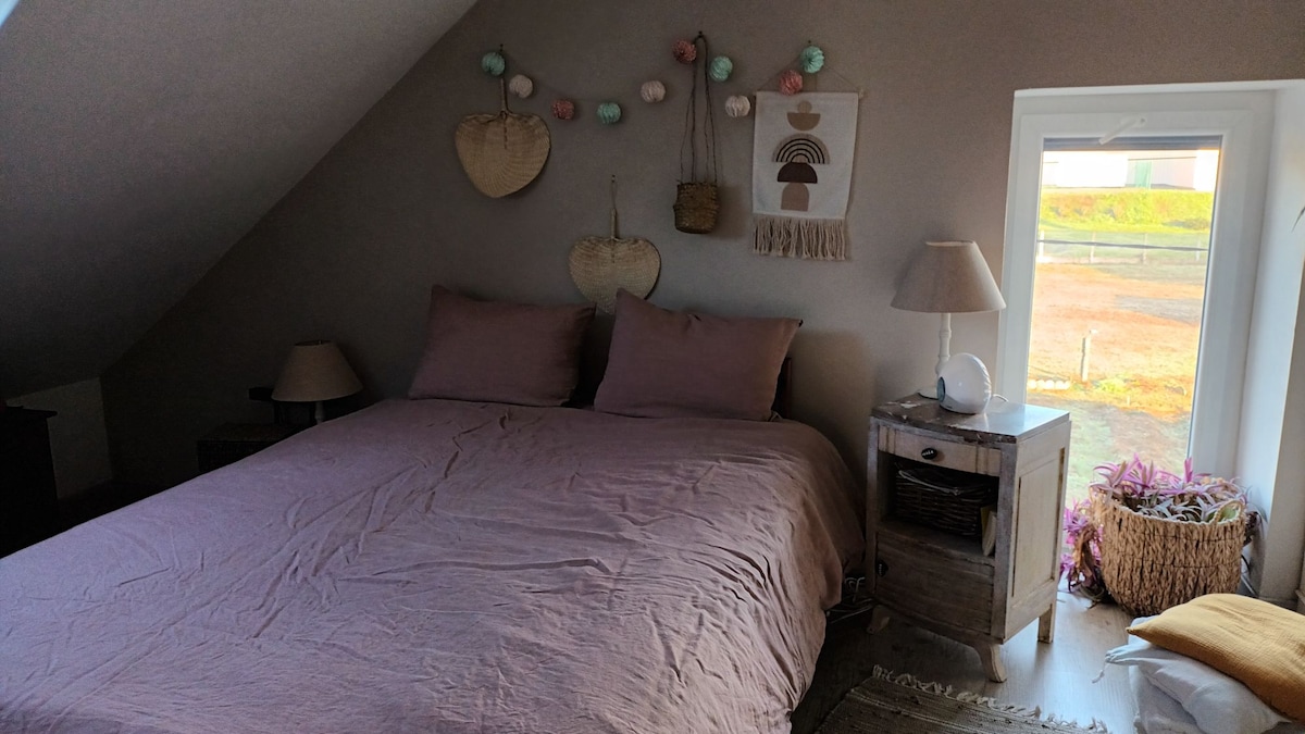 A tranquil bedroom features a spacious bed dressed in soft linens. Decorative wall hangings add subtle texture, while a window allows natural light to fill the space. A bedside lamp and a small nightstand are present, contributing to the cozy setting.