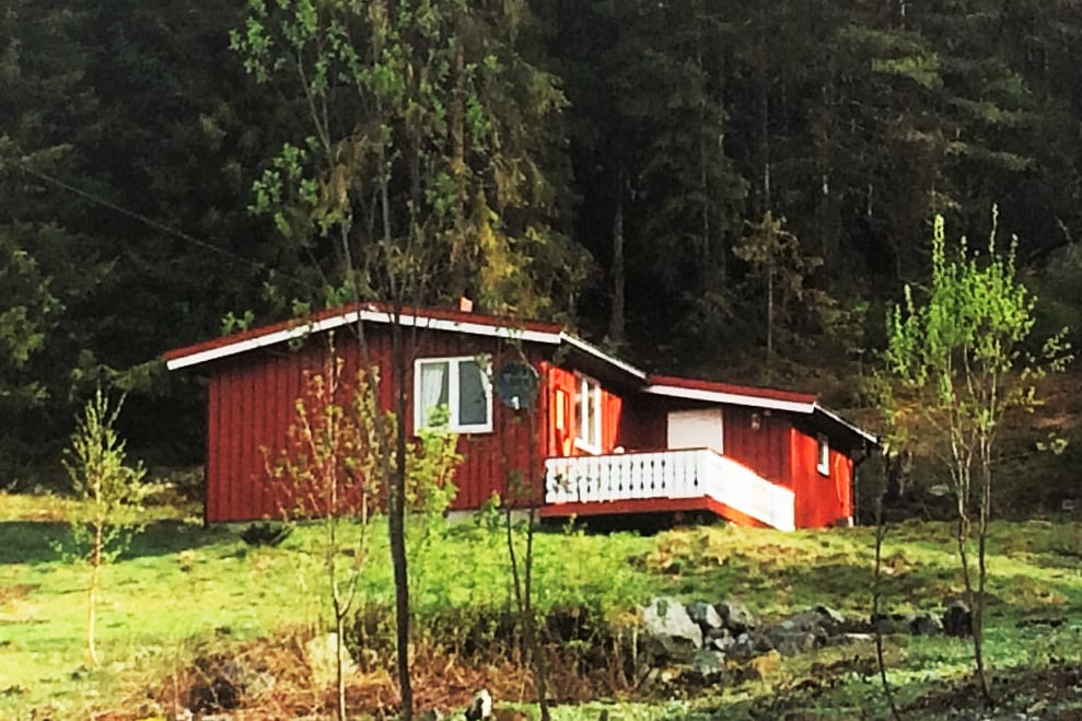 A red cottage is nestled in a quiet forest. Surrounded by greenery, the building features a sloped roof and a porch with a railing. Tall trees provide shade, with a well-kept grassy area visible in the foreground.
