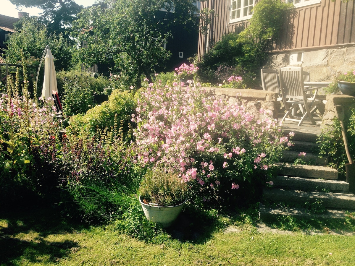 A vibrant garden is presented, featuring an array of blooming flowers in shades of pink and purple. Stone steps lead towards a small seating area with a table and chairs. Lush greenery surrounds the garden, creating a serene outdoor space.