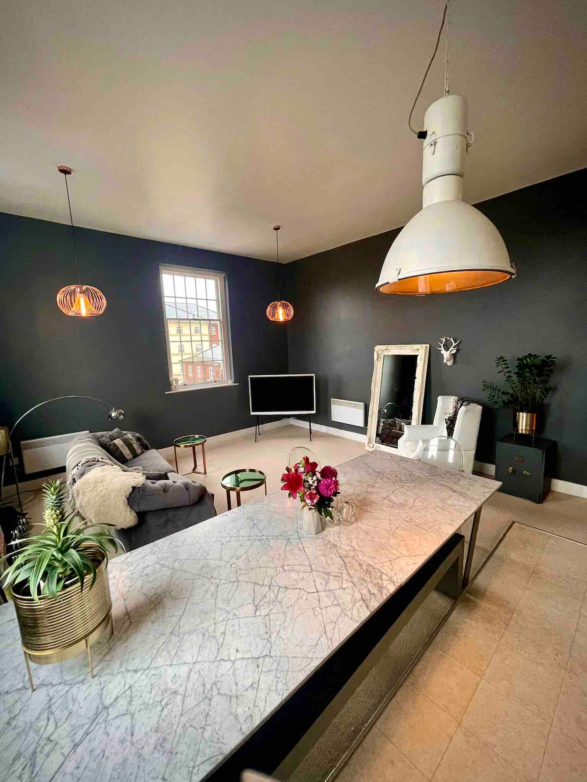 The spacious living area features a marble countertop island, adorned with a small bouquet of flowers. Soft seating is arranged across from a flat-screen television, complemented by stylish lighting fixtures and decorative mirrors, creating an inviting space for relaxation.