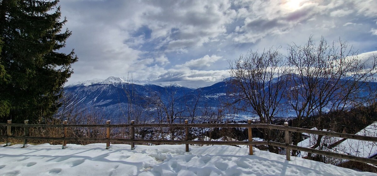A scenic outdoor view showcases snow-covered terrain bordered by a wooden fence. Mountains rise in the distance under a cloudy sky, with patches of blue visible. Leafless trees with branches contrast against the crisp, white snow, conveying a serene winter landscape.