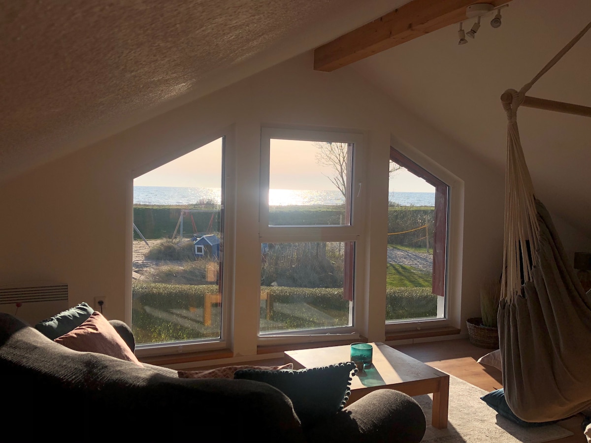 A bright living area features large, triangular windows offering views of the shimmering sea. Natural light floods the space, highlighting a cozy seating arrangement with a dark sofa and a wooden coffee table, creating an inviting atmosphere for relaxation.