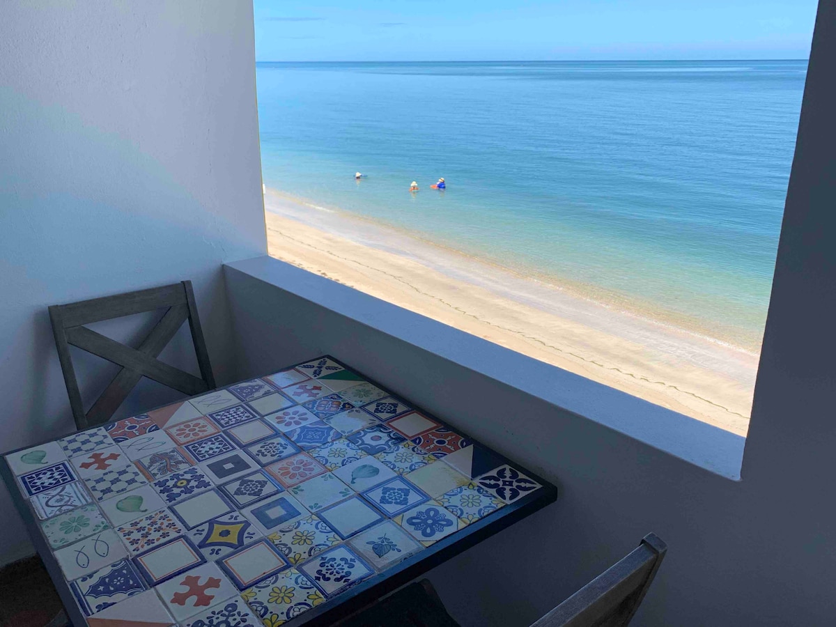 An outdoor seating area features a tiled table with colorful patterns. A clear view of the calm ocean is visible through the open window, with sandy beach and distant beachgoers enjoying the serene surroundings.