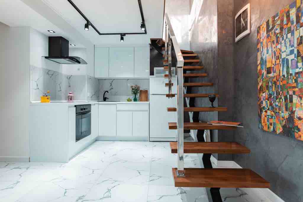The open-concept kitchen features modern cabinetry, a sleek stove, and a marble backsplash. A wooden staircase with a metal railing ascends to the upper level. A striking multicolored artwork is mounted on the wall, adding visual interest to the space.