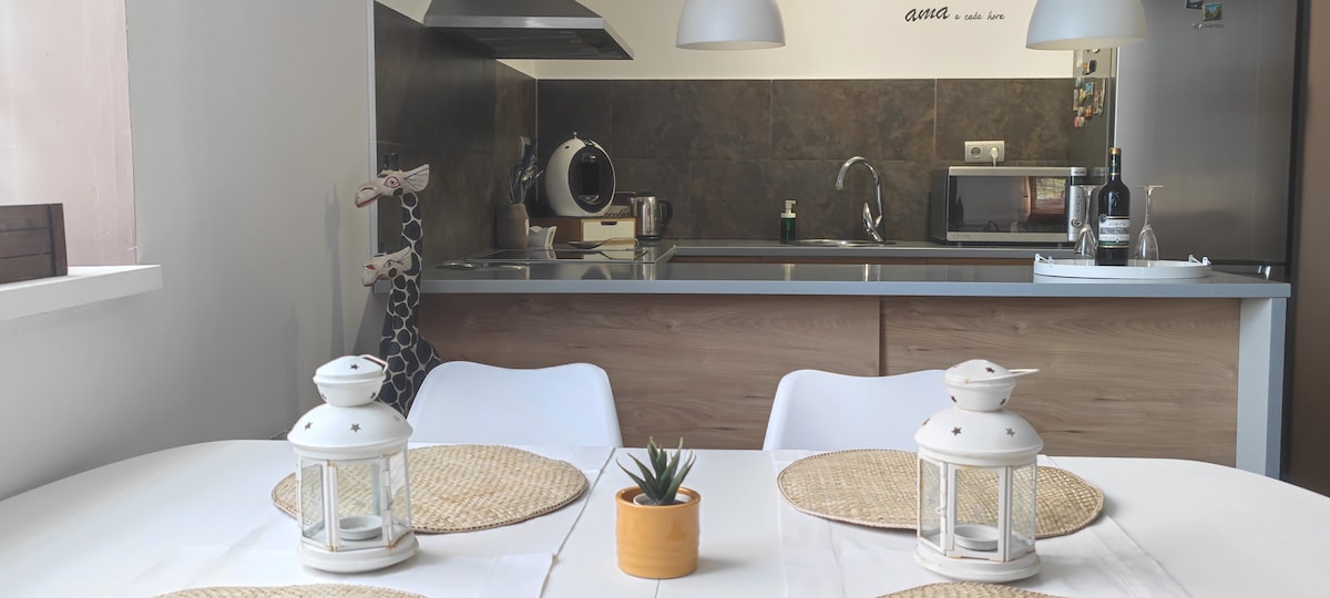 A modern dining area features a round table set with four placemats and lanterns. A small potted plant is centered on the table. The open kitchen is visible in the background, showcasing sleek counters and essential appliances.