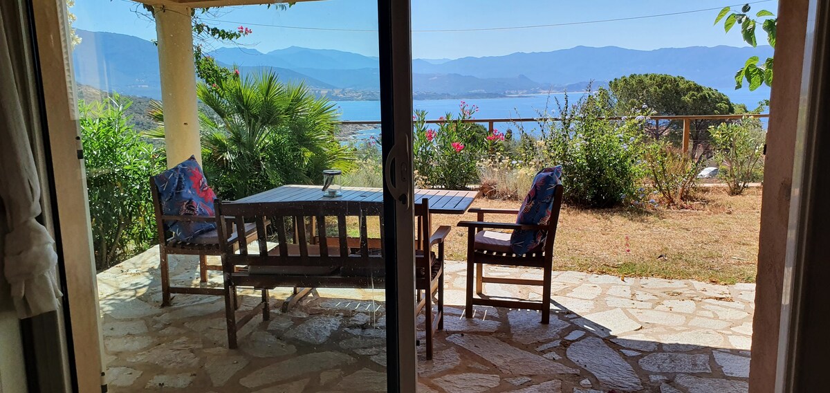 A spacious outdoor terrace is visible, furnished with a wooden table and chairs, surrounded by greenery and flowers. A view of the distant mountains and sea enhances the tranquil environment. Sunlight casts gentle shadows on the stone flooring, particularly inviting for outdoor dining.