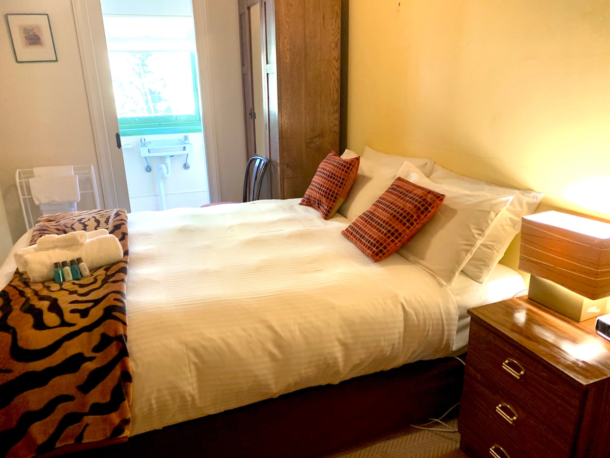 A petite room features a spacious bed dressed in white linens with decorative pillows. A bedside table with a lamp sits next to the bed. A softly lit entrance leads to an ensuite bathroom visible in the background.