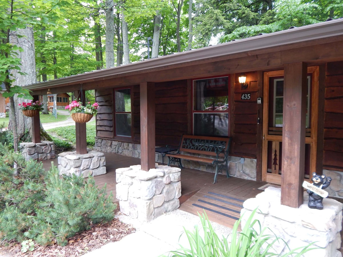 The entrance of the cottage features a covered porch with wooden pillars and stone accents. Two flower baskets hang at either end, adding natural color. A rustic bench is positioned beside a warm wooden door, set against a backdrop of lush green trees.