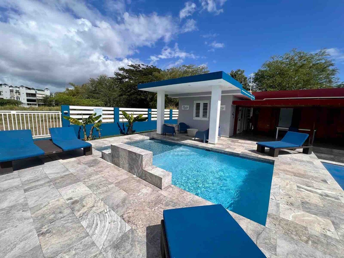 A modern pool area is showcased, featuring a rectangular pool surrounded by stylish stone tiles. Comfortable lounge chairs in a deep blue hue are placed on one side. A shaded outdoor structure with a blue roof offers seating and a relaxing atmosphere. Lush greenery is visible in the background.