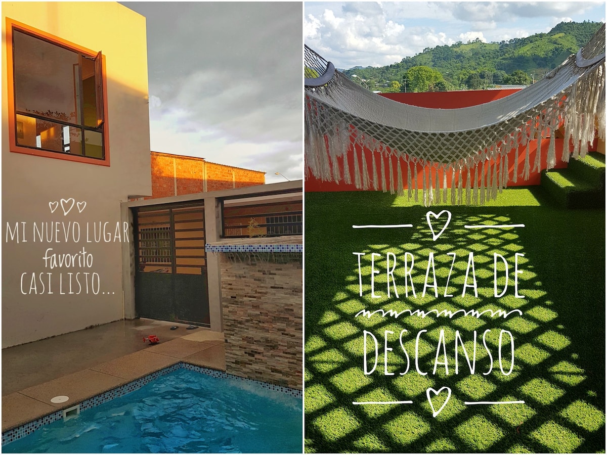 A refreshing swimming pool is visible, bordered by textured stone walls. Adjacent, a serene terrace with synthetic grass features comfortable hammocks, surrounded by natural plants. The area offers views of the lush green mountains in the distance, creating an inviting outdoor space.