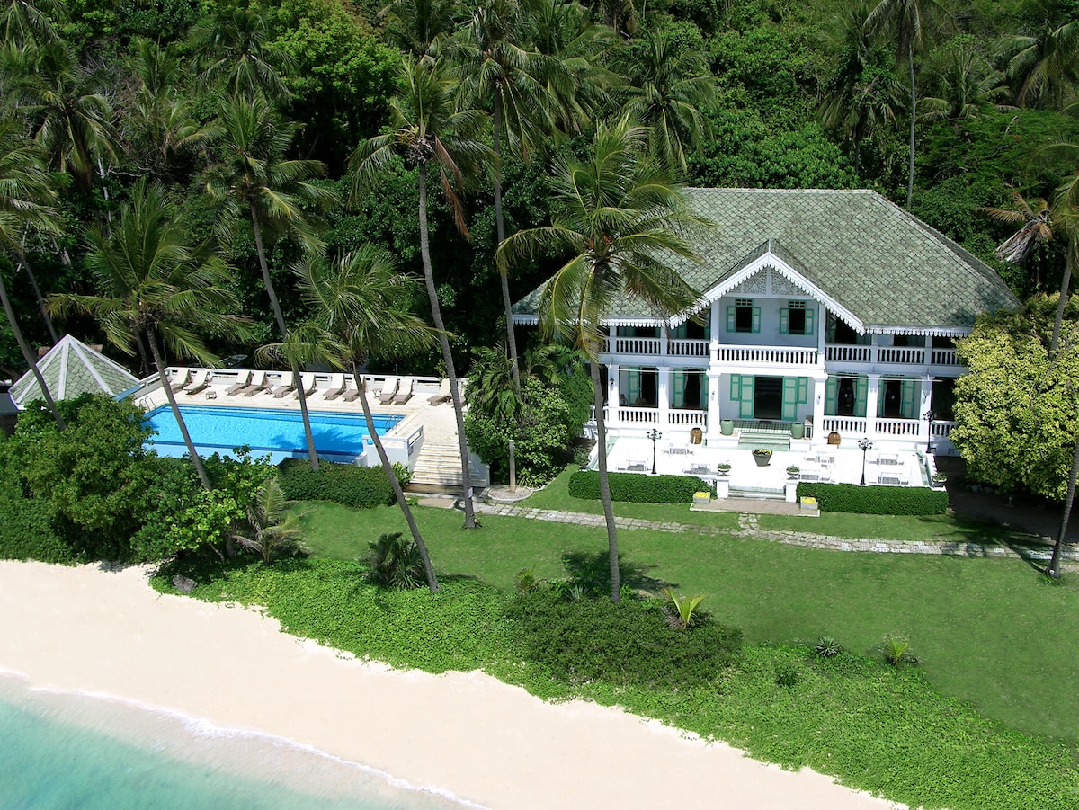 A spacious villa is shown with a tiled roof, set amid lush greenery and swaying palm trees. A long swimming pool is visible in front of the villa, accompanied by sun loungers. The property is situated near a sandy beach, enhancing the tranquil tropical ambiance.