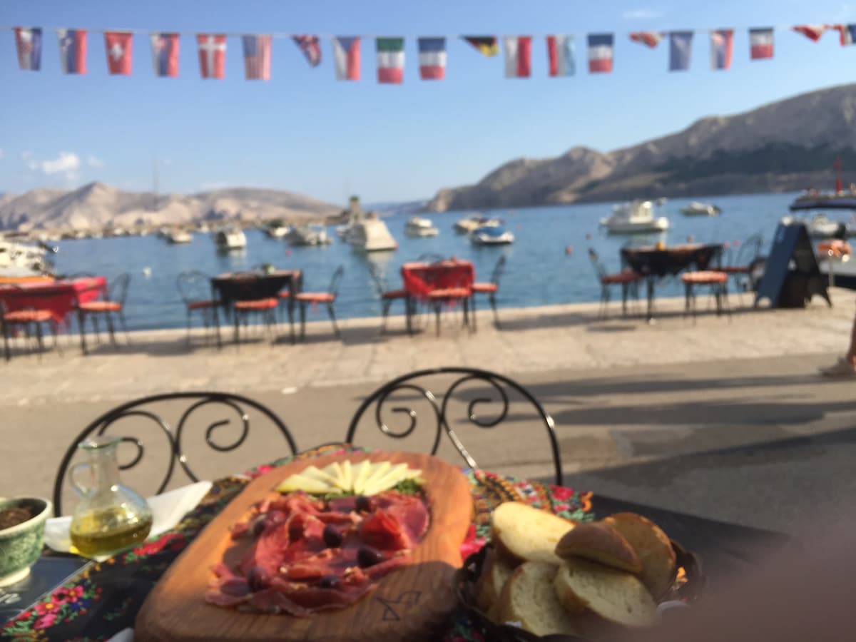 A wooden platter is displayed featuring assorted cured meats and slices of cheese, accompanied by fresh bread. A picturesque waterfront scene is visible in the background, with several boats anchored and mountains in the distance. Colorful flags are strung along the shoreline.
