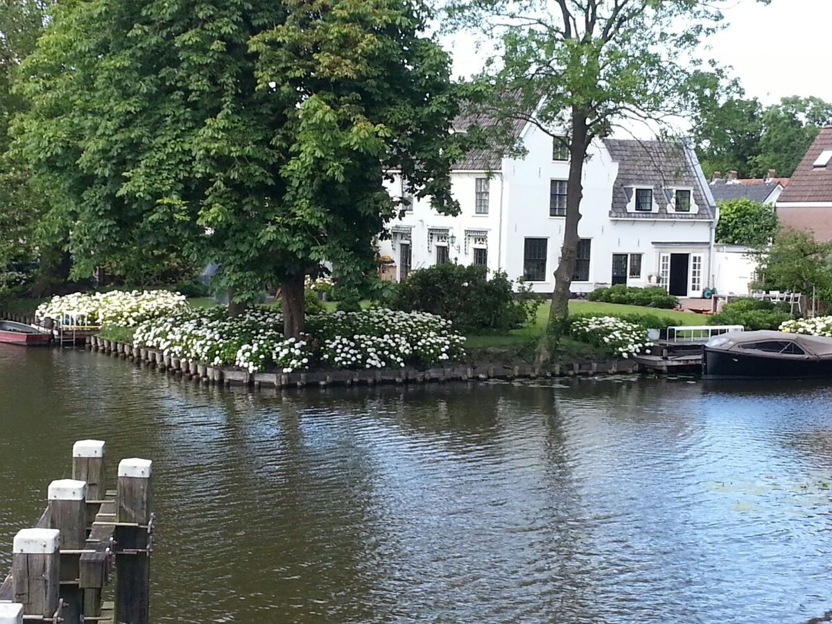 A charming view of a white two-story house is set against a backdrop of lush greenery and flowering plants. The tranquil water reflects the surrounding landscape, while a small jetty is visible in the foreground. The scene captures the peaceful essence of riverside living.
