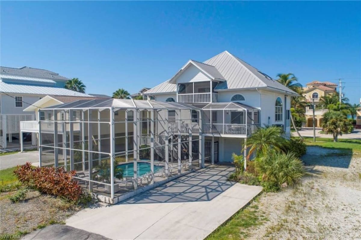 The two-story home is surrounded by a lush landscape, featuring a screened-in area that houses a private pool. A spacious patio area with seating is visible, inviting relaxation. The exterior is painted in soft tones, with a gabled roof and multiple balconies accessible from the upper level.