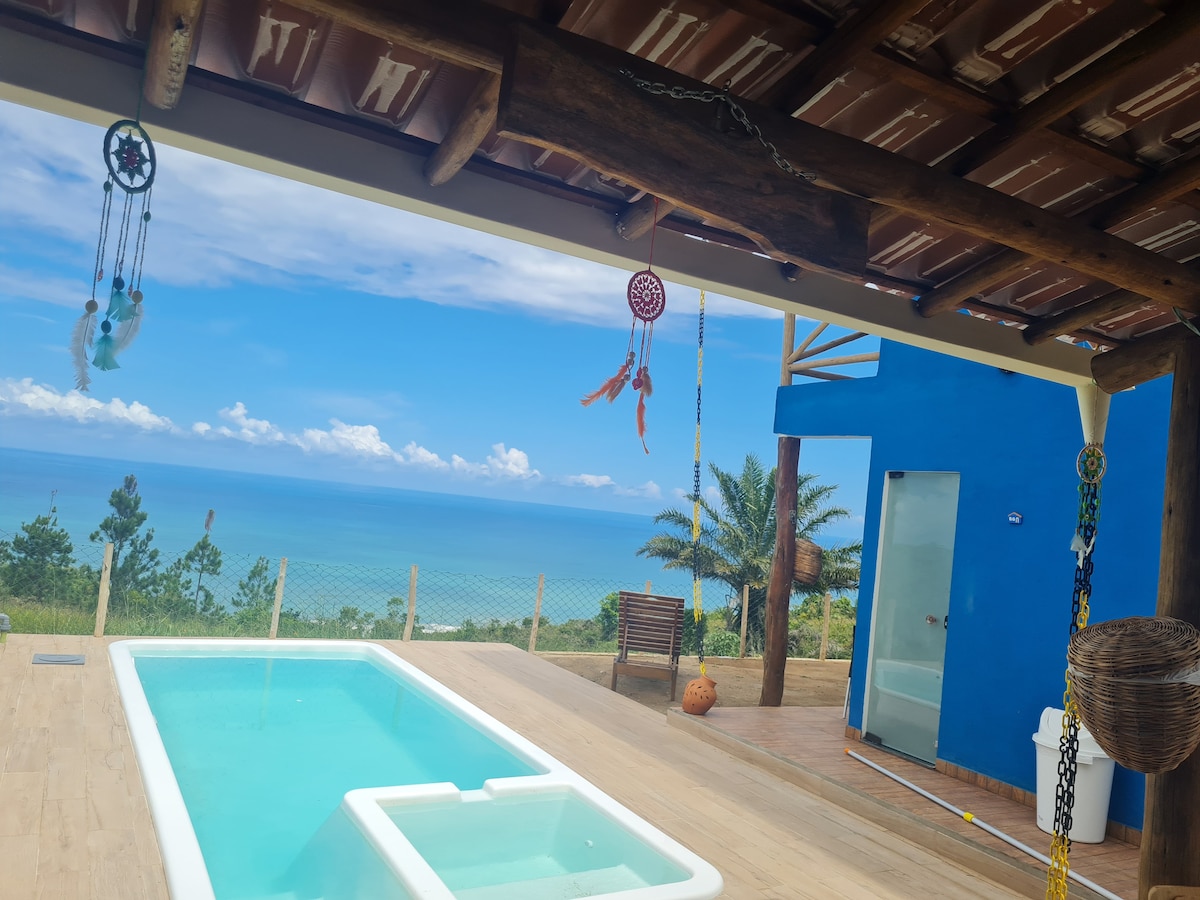 An outdoor space features a private pool adorned with colorful dreamcatchers and a view of the sea in the background. The area is framed by wooden beams and vibrant blue walls, providing a tranquil retreat amidst nature.