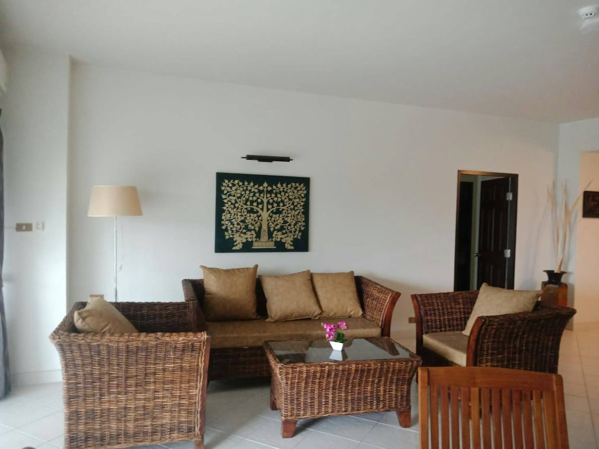 A cozy seating area is arranged with woven wicker furniture, featuring a brown sofa with plush cushions and two matching armchairs. A glass-topped coffee table sits in the center, accompanied by a small vase of flowers. A decorative wall hanging adds a touch of style.