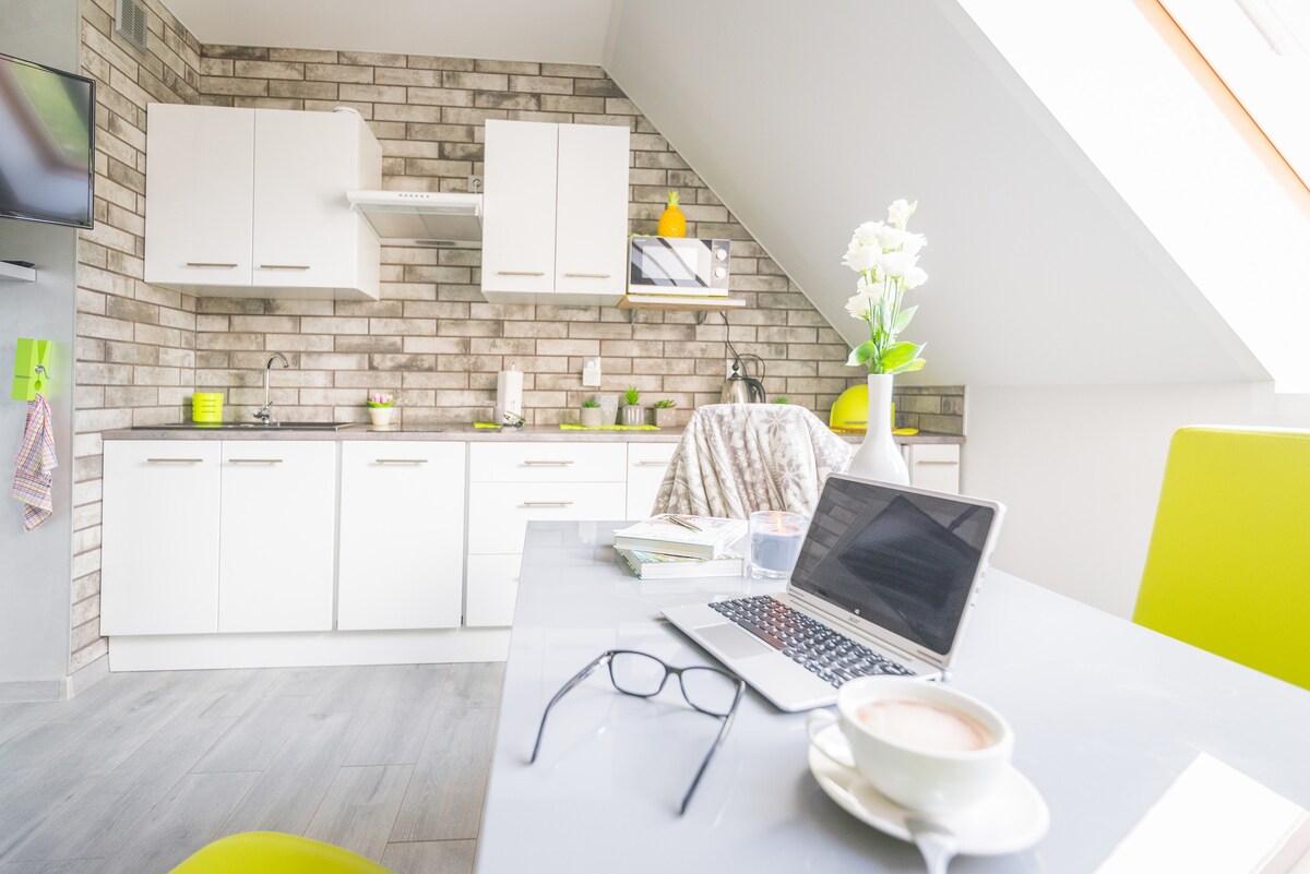 The kitchen area features modern cabinetry with a minimalist design, complemented by a gray tiled wall. A dining table is set with a laptop, glasses, and a cup, while fresh flowers add a touch of color. Sunlight streams through the angled window, enhancing the space.