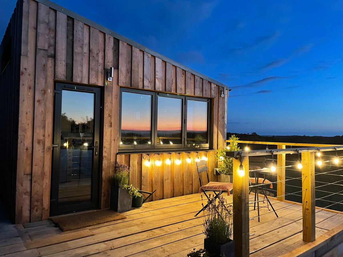 The cabin exterior is showcased under a twilight sky, featuring wooden paneling and large windows. A deck with string lights and bistro furniture creates an inviting space to enjoy the sunset view. The serene ambiance is enhanced by the surrounding nature.