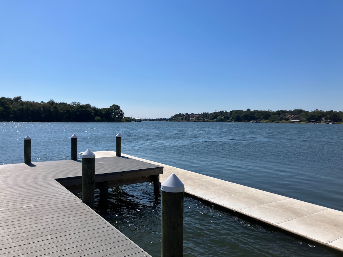 A view of a calm waterfront scene is captured from a wooden dock, featuring sturdy posts topped with white caps. The serene water reflects the clear blue sky, while green vegetation borders the distant shore, offering a peaceful atmosphere.